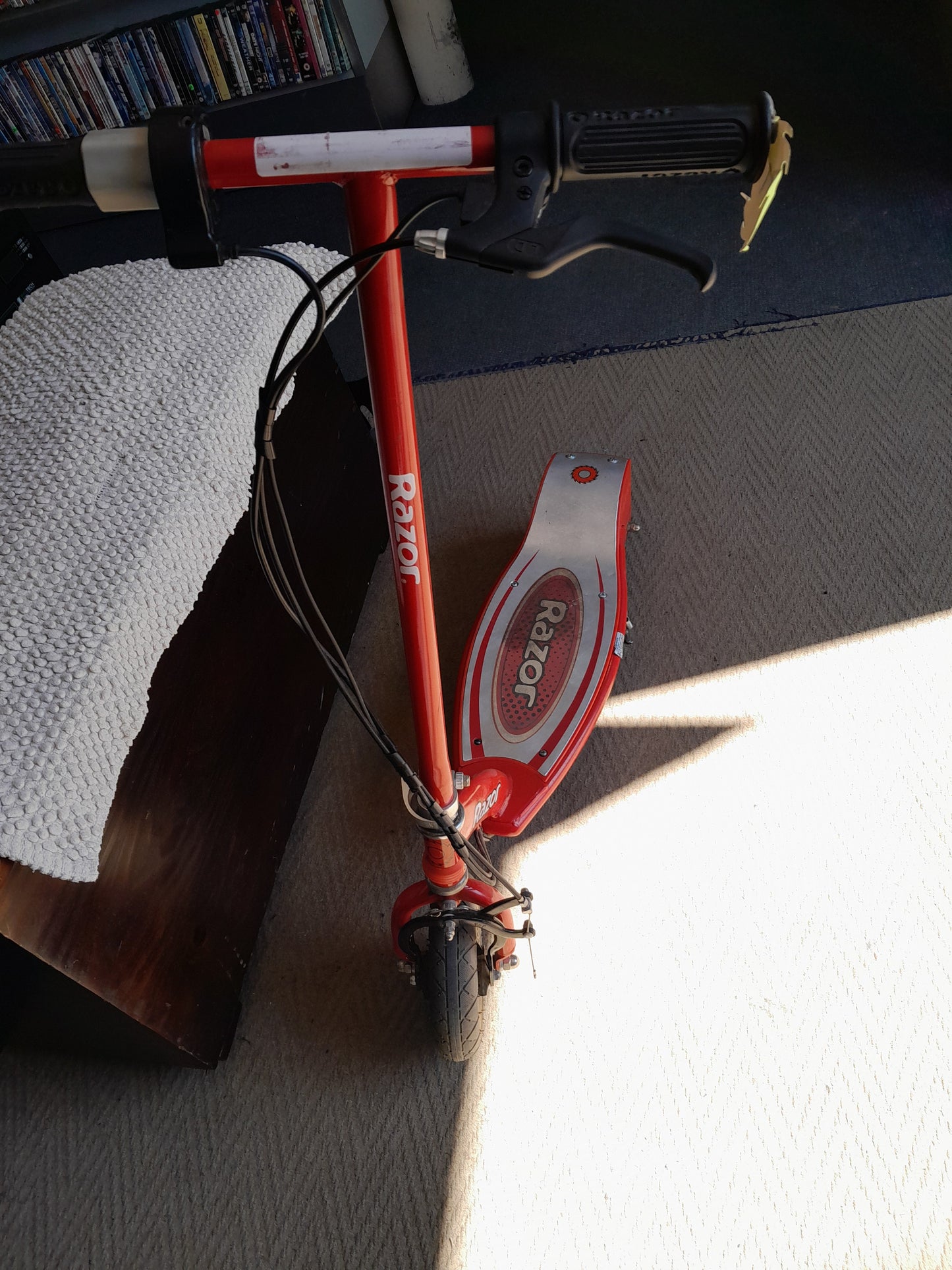 Red Razor electric scooter positioned indoors on a carpeted floor with visible handlebar, brake lever, and front wheel, available at Die Skatkis.