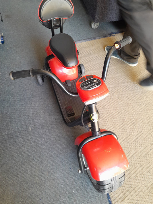 Red 3-wheel electric scooter for kids with a 12V battery and comfortable seat, displayed on a carpeted floor, available at Die Skatkis.