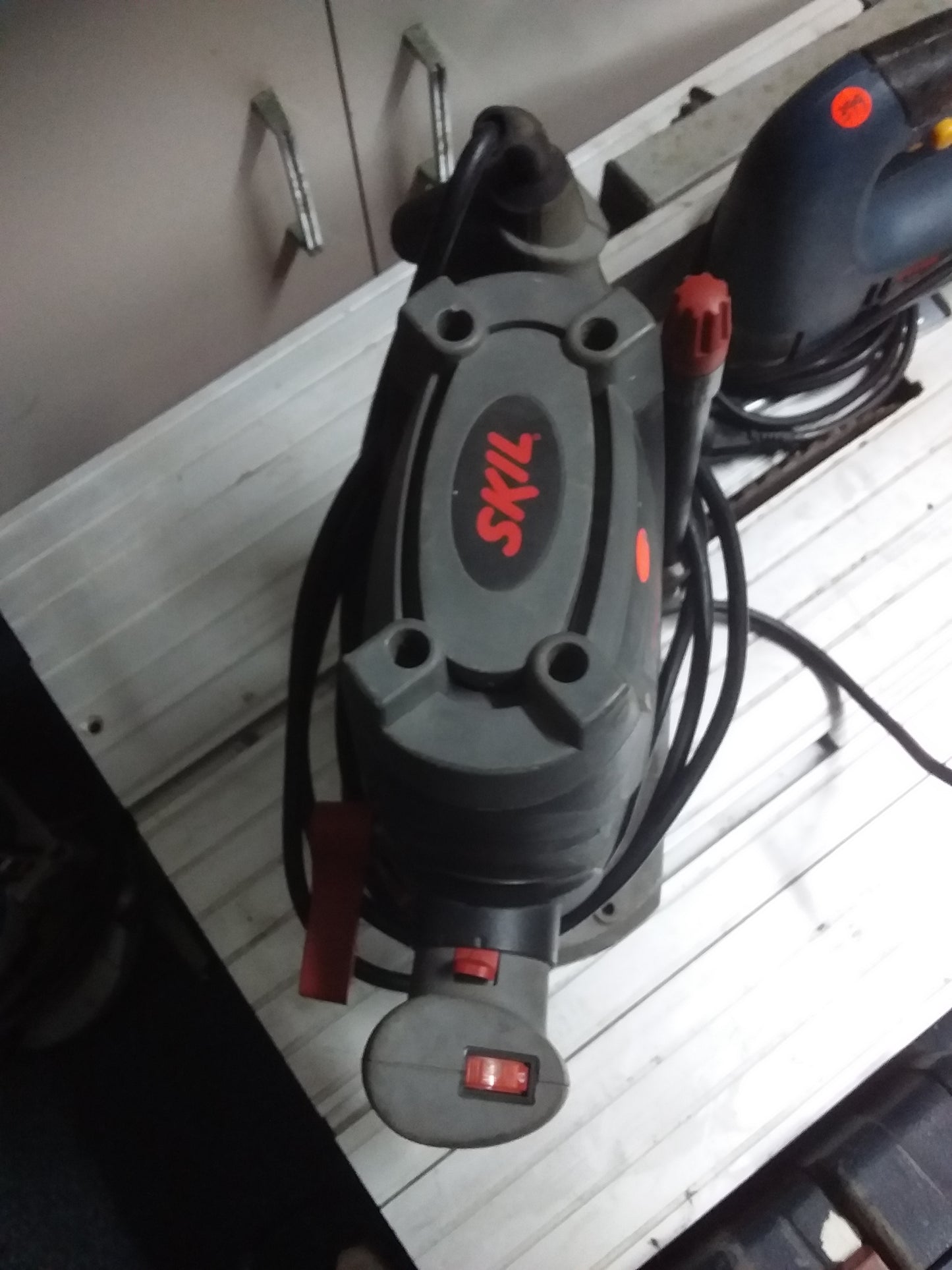 "SKIL Router with red handles and power cords, placed on a white workbench with tools in the background. Available at Die Skatkis.