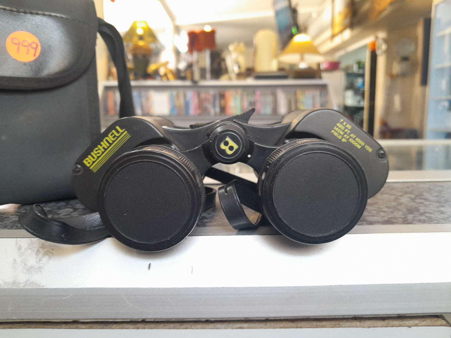 Close-up of Bushnell 7x35 binoculars with lens caps on, text showing 420 FT. AT 1000 YDS. 140 M AT 1000 M FIELD 6º, available at Die Skatkis.