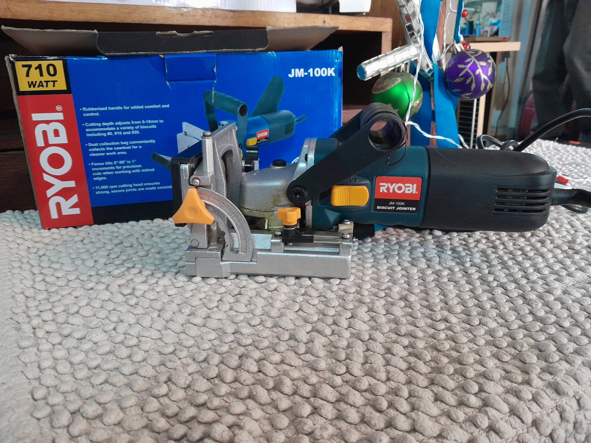Ryobi JM-100K Biscuit Jointer on a textured surface with packaging box highlighting features like 710W motor, adjustable depth.