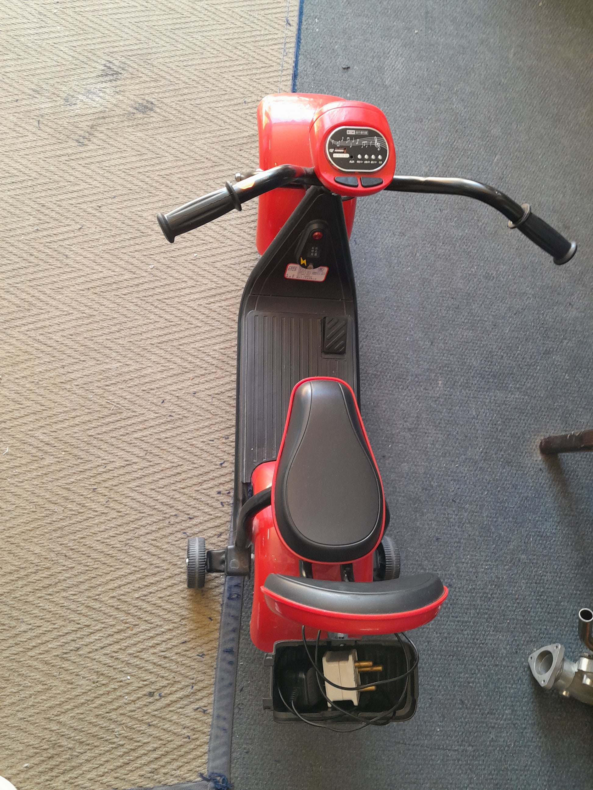 Red and black 3-wheel electric scooter with a seat and handlebar grips, featuring a small storage compartment and white electrical plug, available at Die Skatkis.