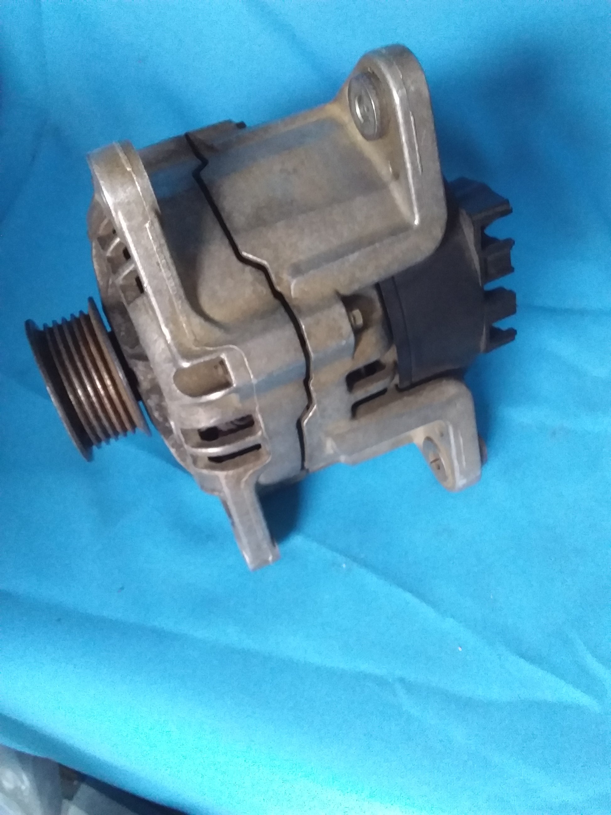 Universal Ford alternator with pulley and electrical connectors, placed on a blue fabric background. Available at Die Skatkis.